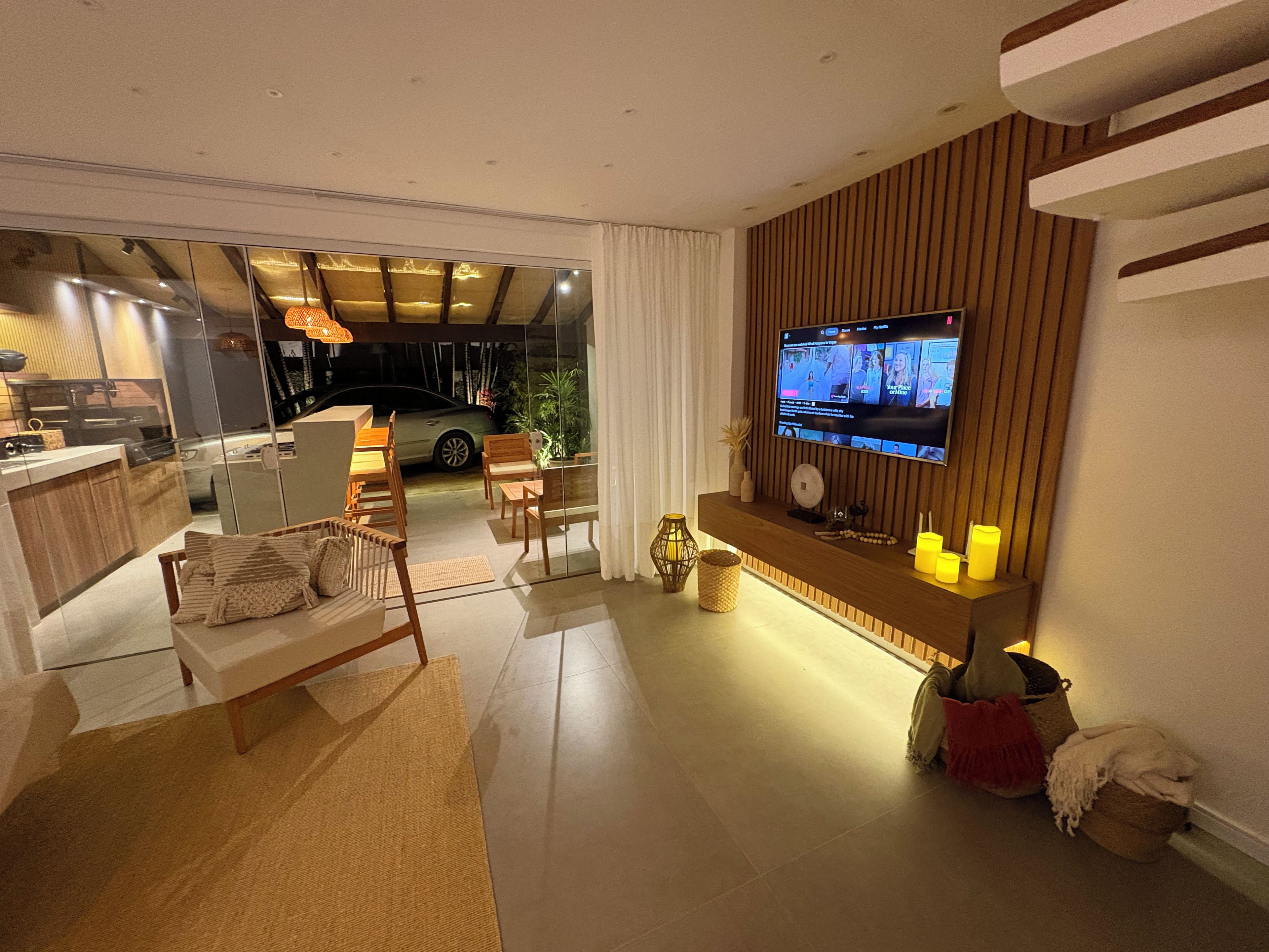 Sala de estar moderna com televisão de tela plana fixada em parede de madeira, área de estar com sofá e poltronas, e vista para um carro através de uma porta de vidro.