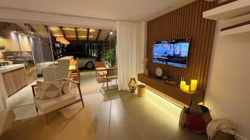 Sala de estar moderna com televisão de tela plana fixada em parede de madeira, área de estar com sofá e poltronas, e vista para um carro através de uma porta de vidro.