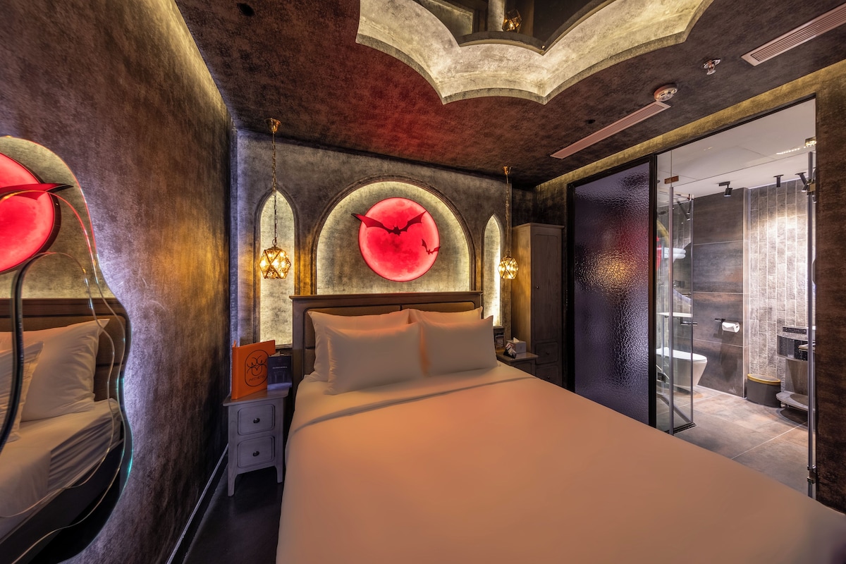 Une chambre d’hôtel moderne avec un grand lit, une table de chevet, une horloge murale et une salle de bain visible à travers une porte en verre.