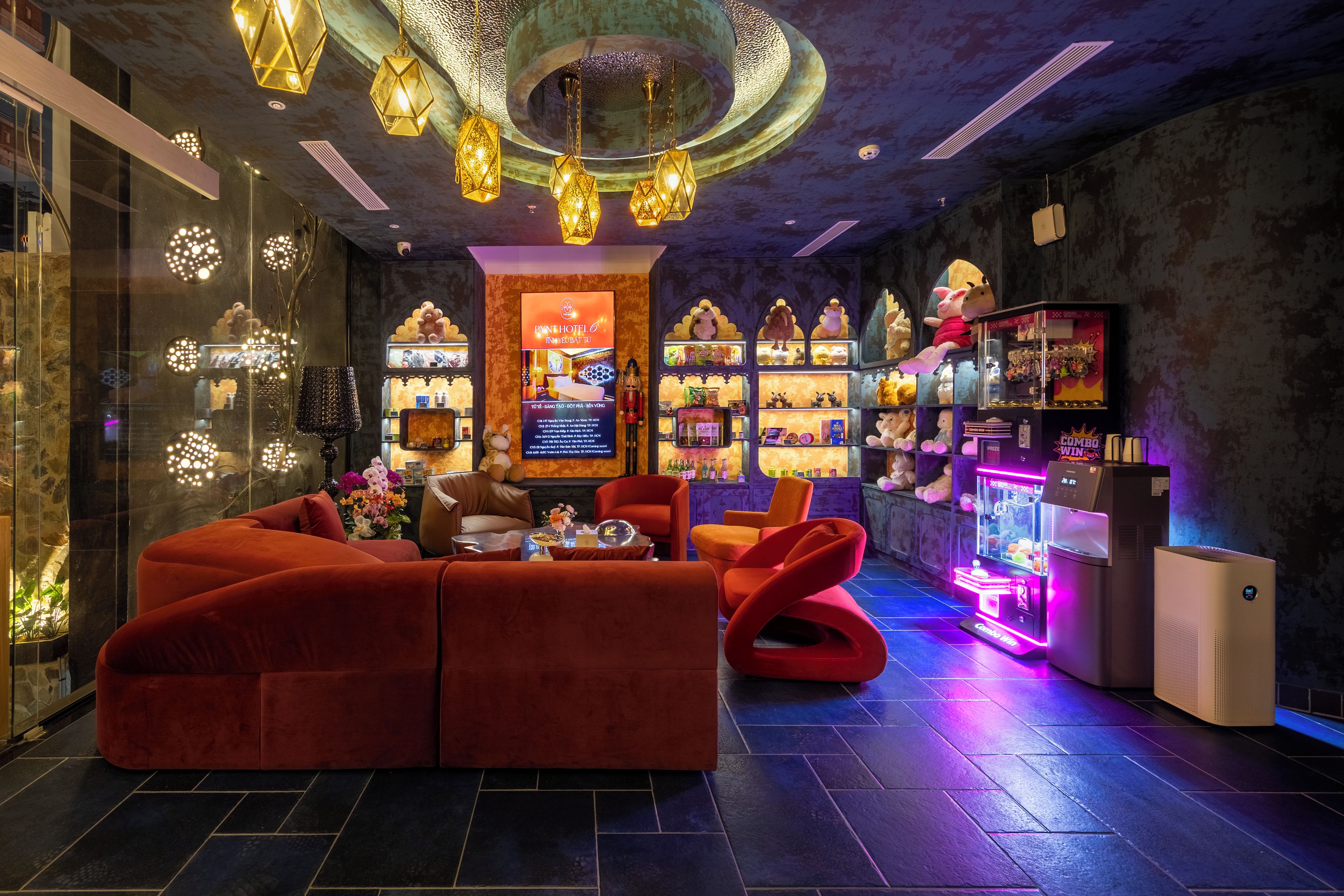 A modern hotel lobby with a plush red sofa, a bar area with claw machines, and a large illuminated sign.