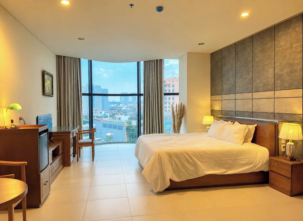 A hotel room with a large bed, a desk, a chair, and a view of the city through large windows.