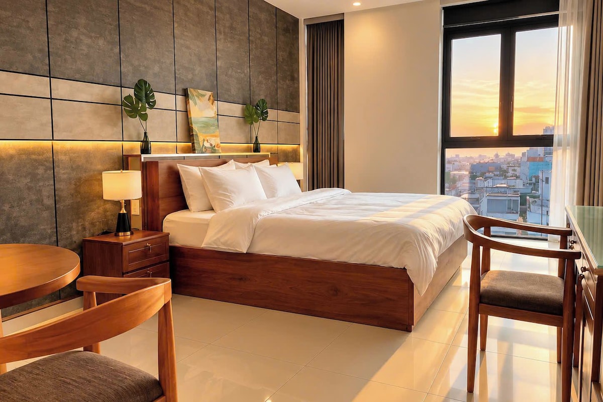 Quarto de hotel com cama, mesa de cabeceira, abajur, cadeira e vista da cidade por uma grande janela.