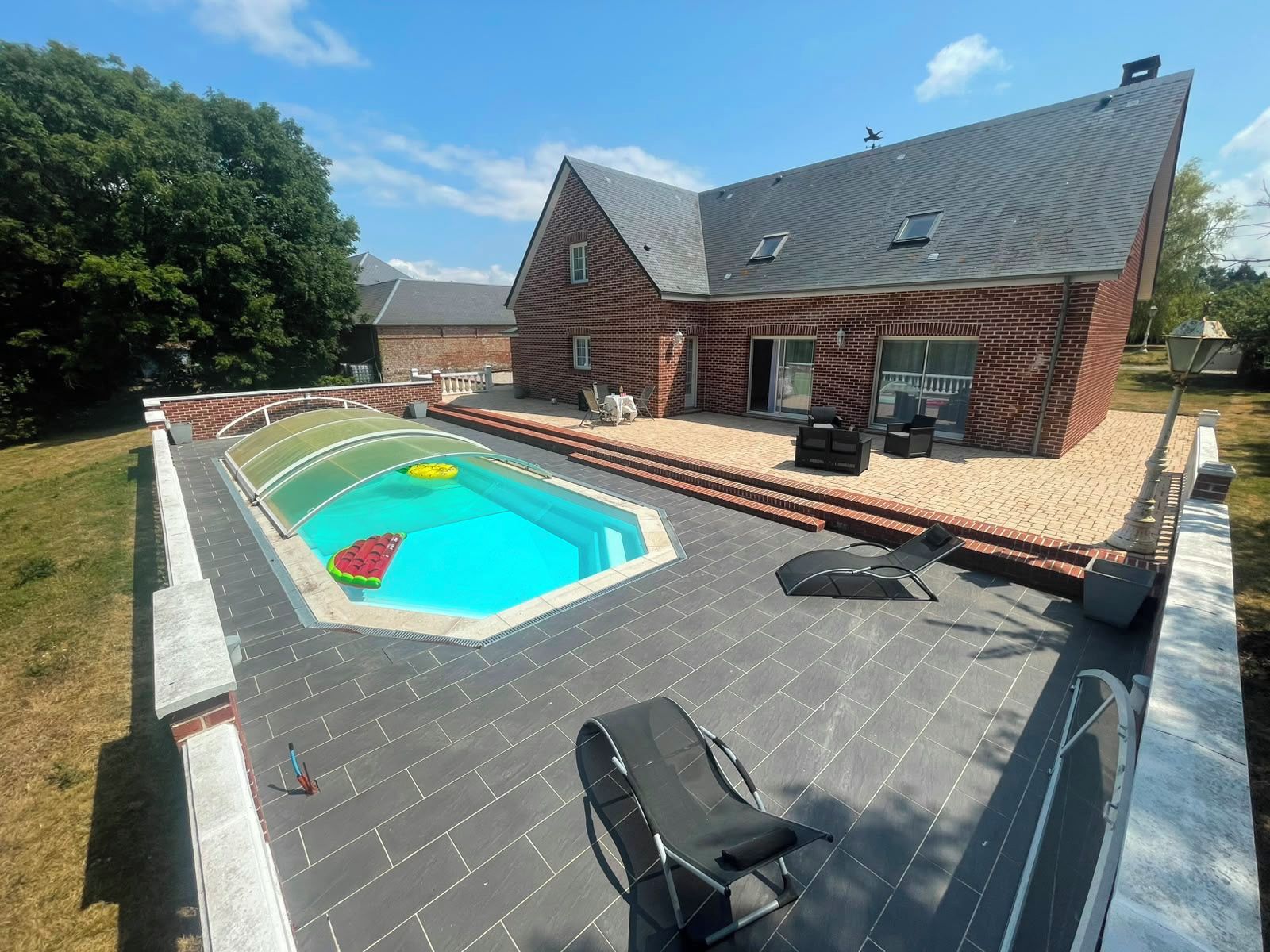A swimming pool with a diving board and lounge chairs, surrounded by a brick house with a sloped roof.