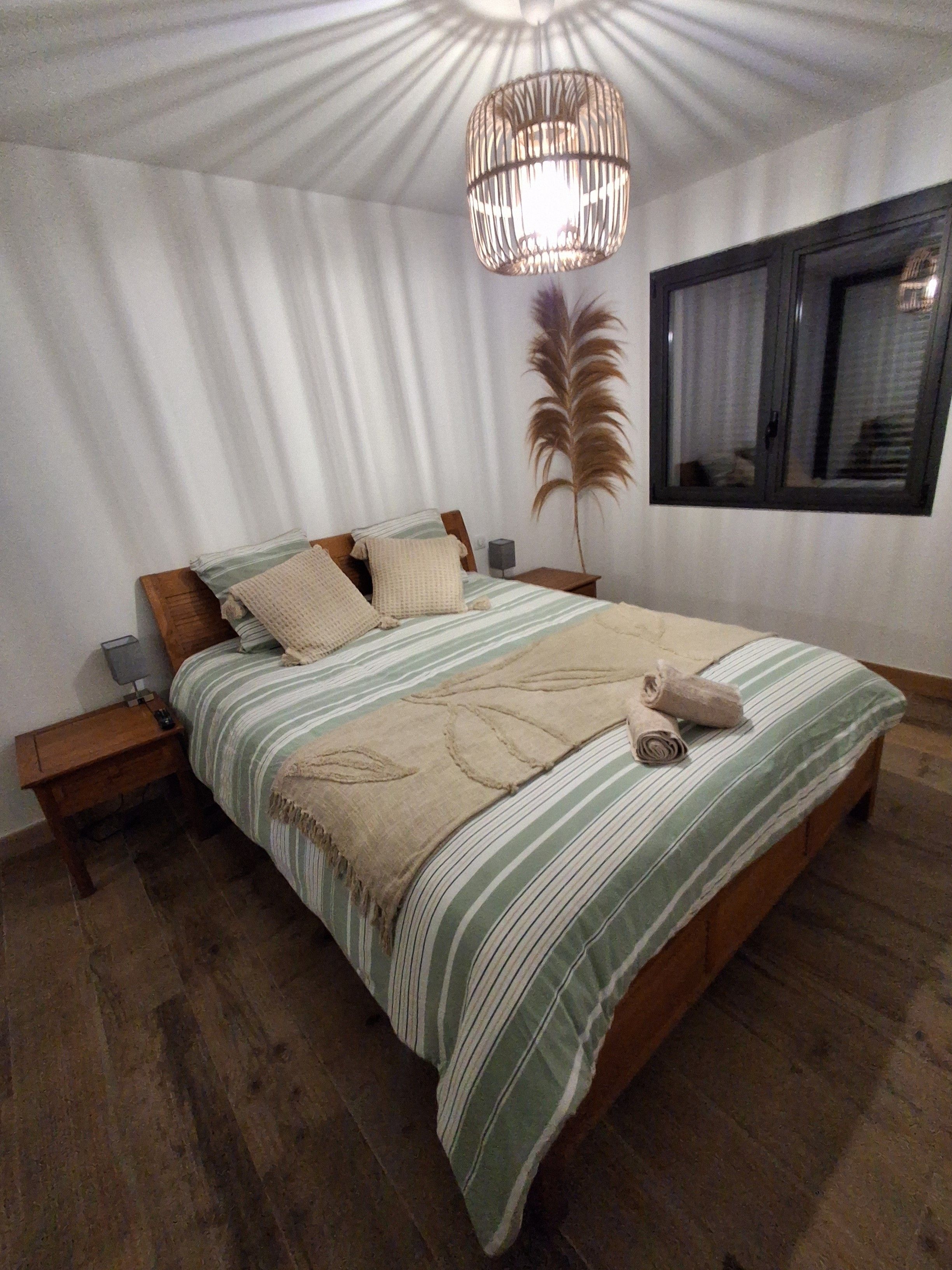 Une chambre à coucher avec un grand lit, une table de chevet, une lampe, une fenêtre et une plante décorative.