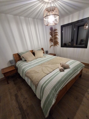 A bedroom with a large bed, a bedside table, a lamp, a window, and a decorative plant.