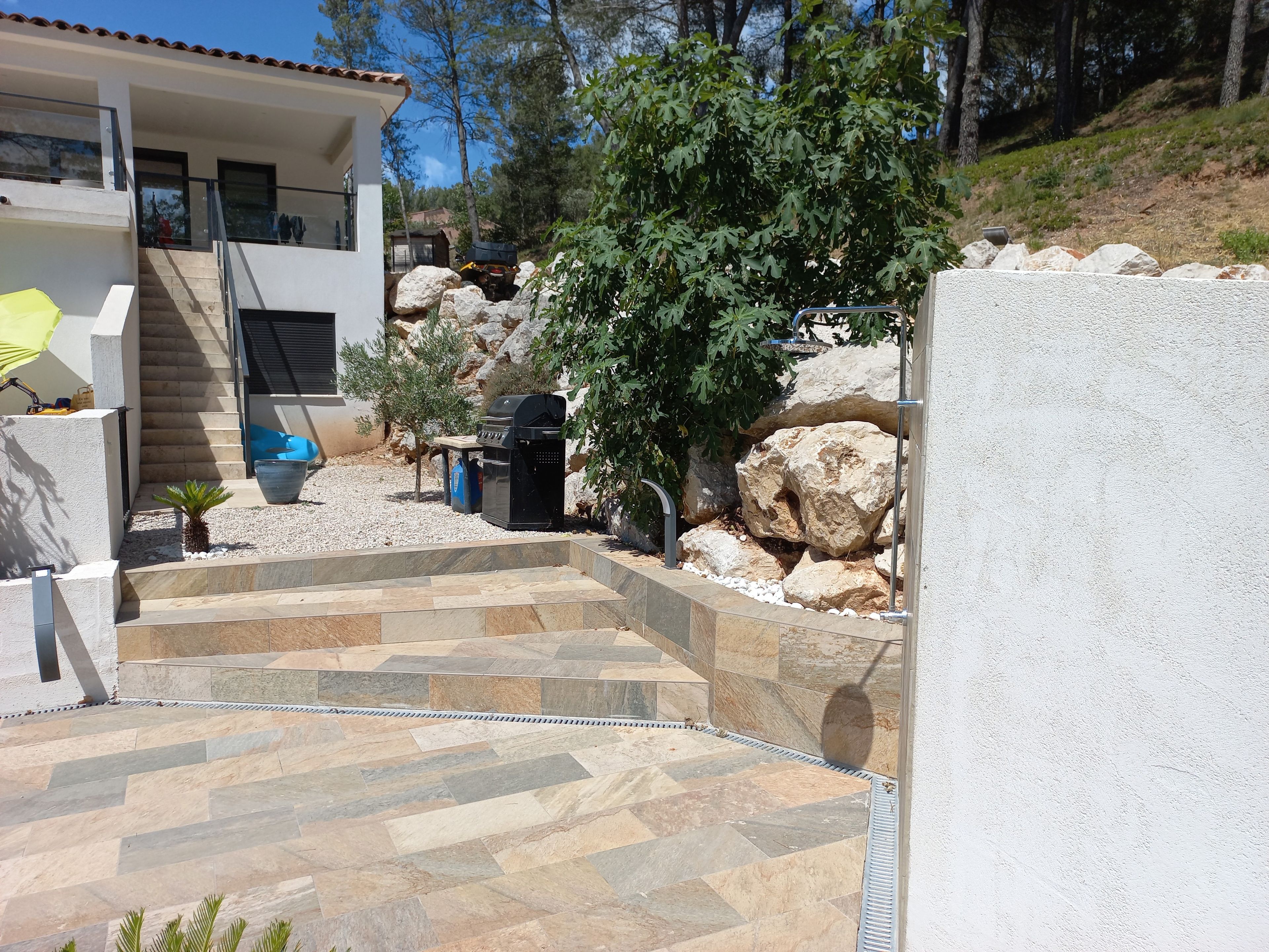 A modern outdoor area with a stone staircase leading to a house, a barbecue grill, and a small palm tree.