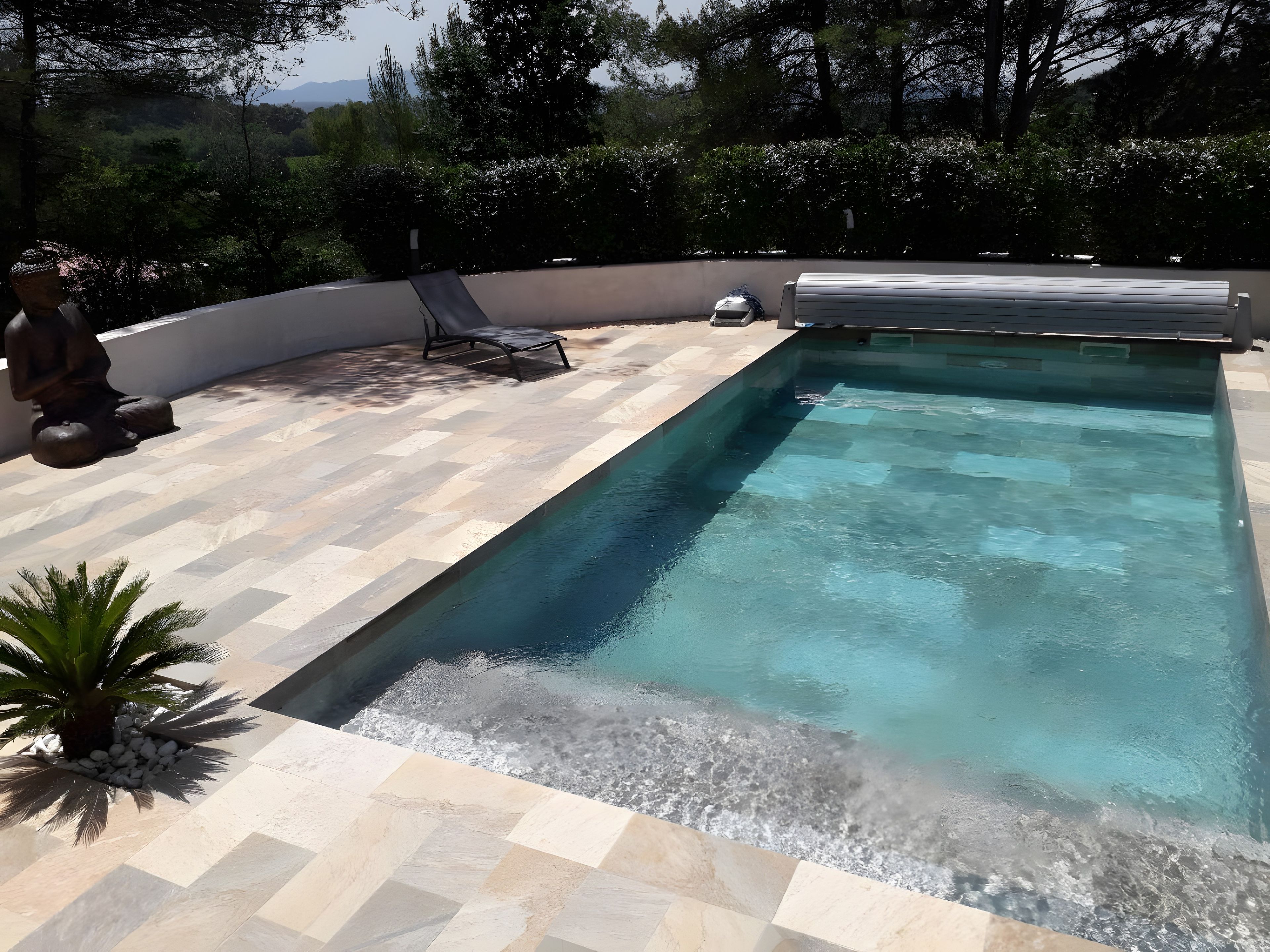 Une piscine rectangulaire avec une terrasse carrelée, un fauteuil de relaxation et un petit palmier.