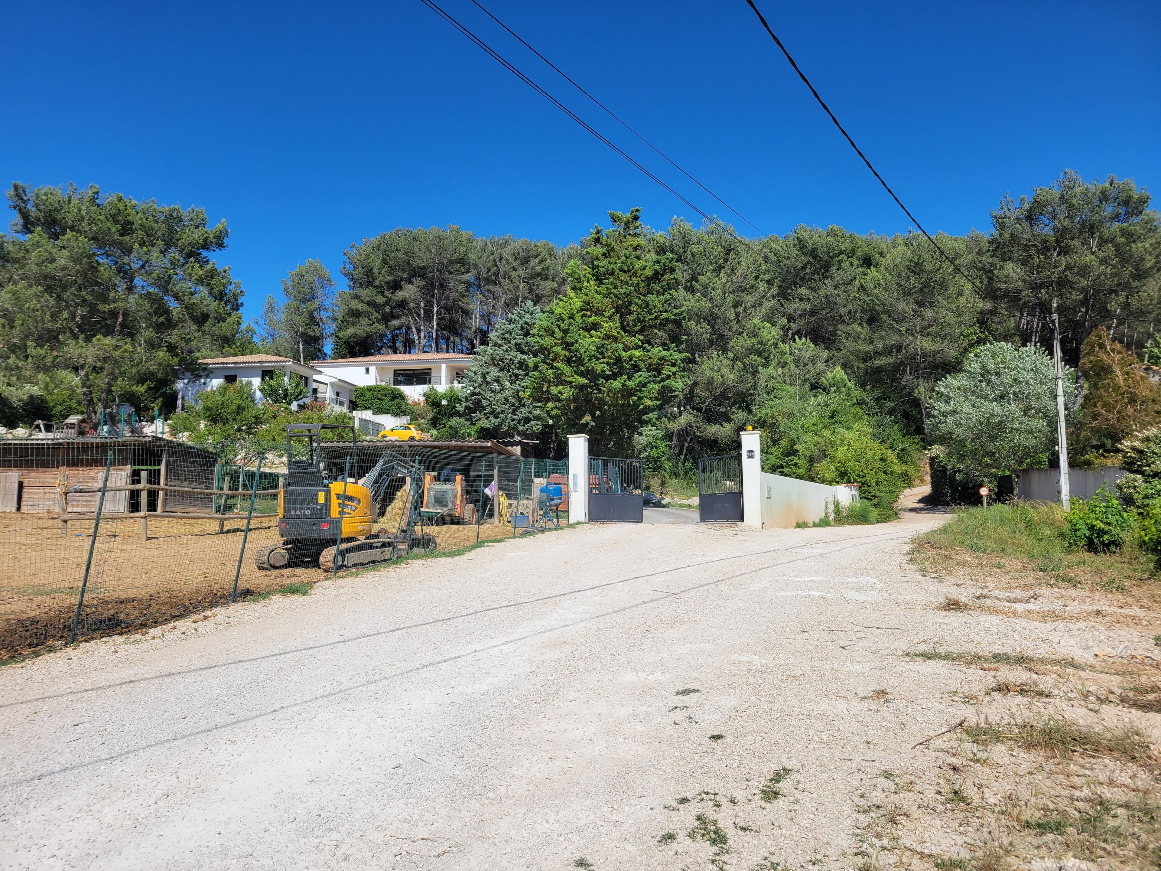 Un chemin de gravier menant à une zone fermée par une barrière, où se trouvent des engins de chantier et un enclos délimité par une clôture.