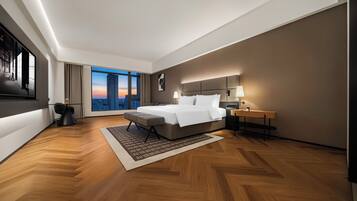 A modern hotel room with a large bed, a desk, a chair, a nightstand, and a view of the city at sunset.