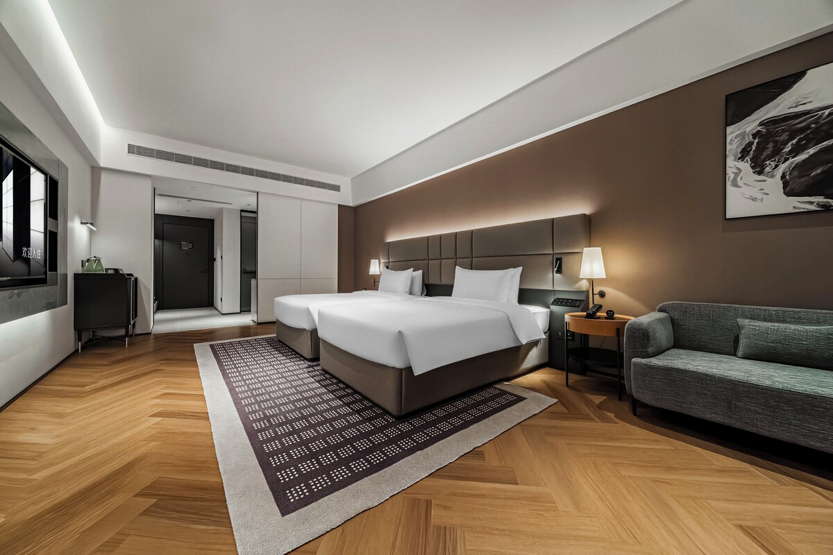 A modern hotel room with a large bed, a sofa, a desk, and a painting on the wall.