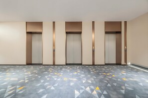 A hallway with five elevators, each with a control panel on the wall. The floor has a geometric pattern.