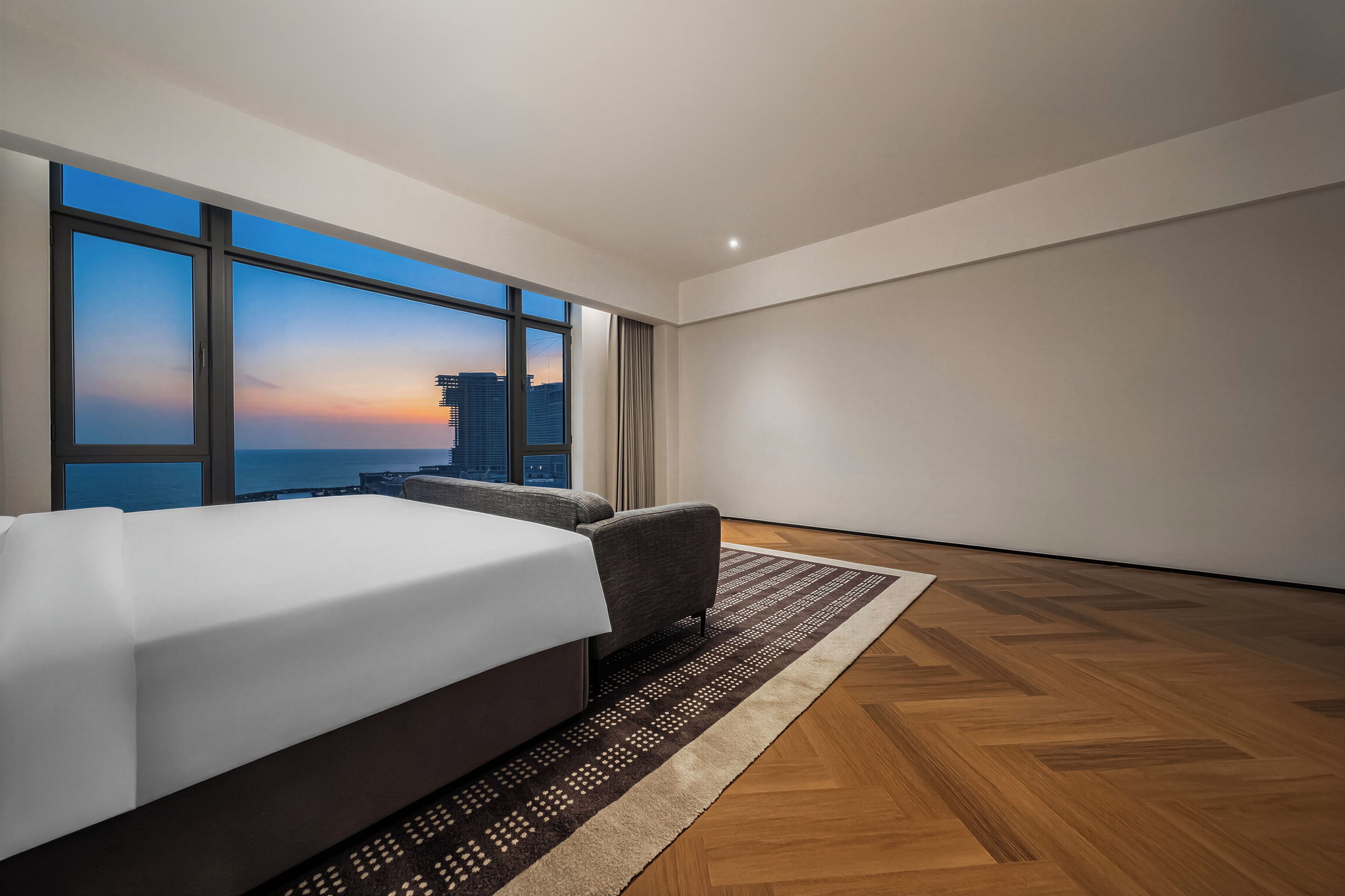 Ein modernes Hotelzimmer mit einem großen Bett, einer Sitzecke und Blick auf die Stadt durch große Fenster.