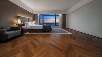 A hotel room with a bed, a sofa, a nightstand, a lamp, and a view of the sunset over the ocean.