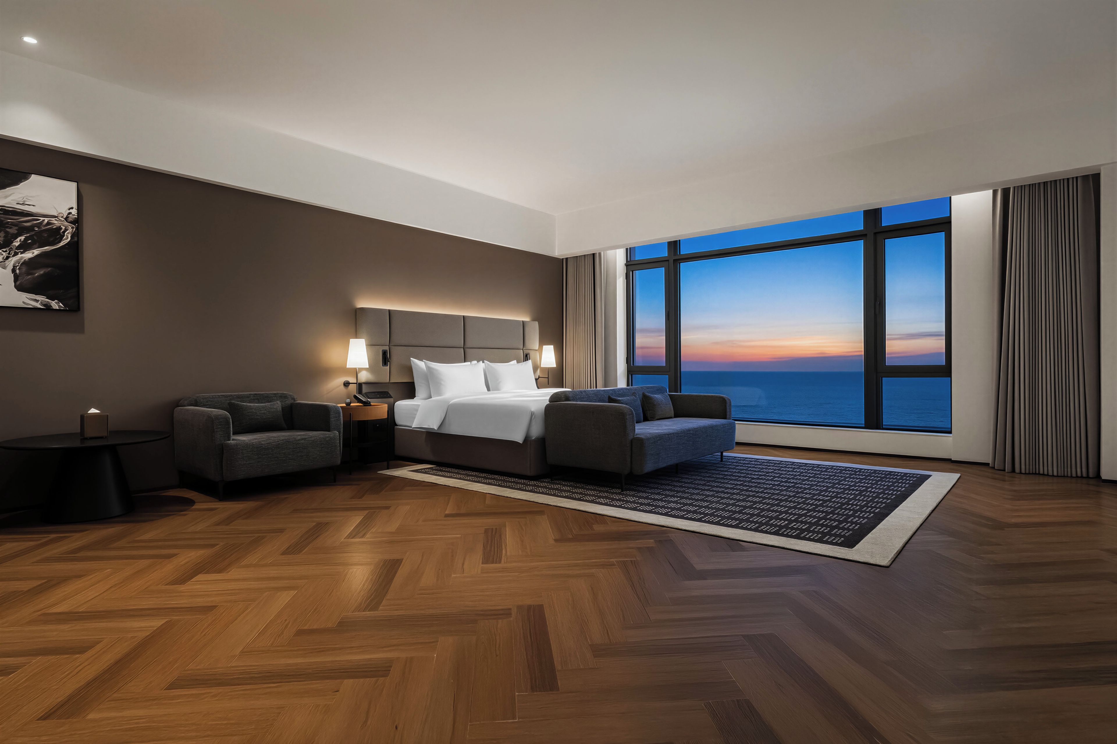 A hotel room with a large bed, two armchairs, a nightstand, and a view of the ocean at sunset.