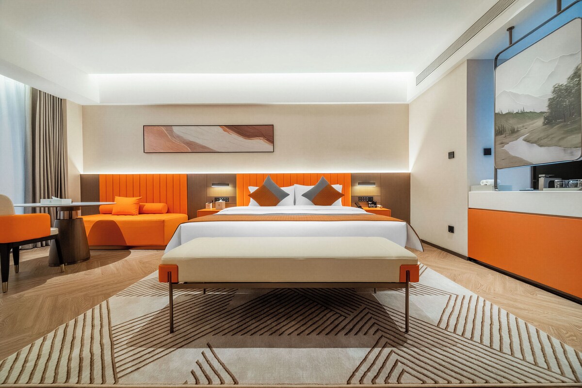 A modern hotel room with a large bed, orange accents, a painting above the bed, and a view of a landscape through a window.