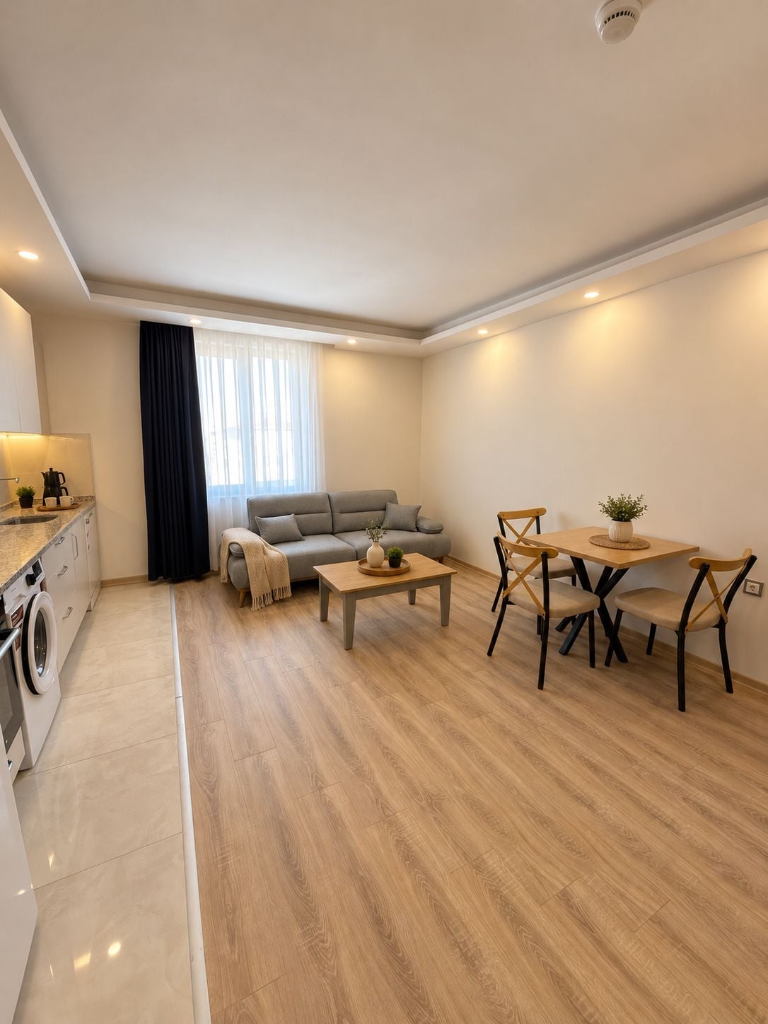 A modern living room with a sofa, coffee table, and dining table. There is a washing machine in the corner.