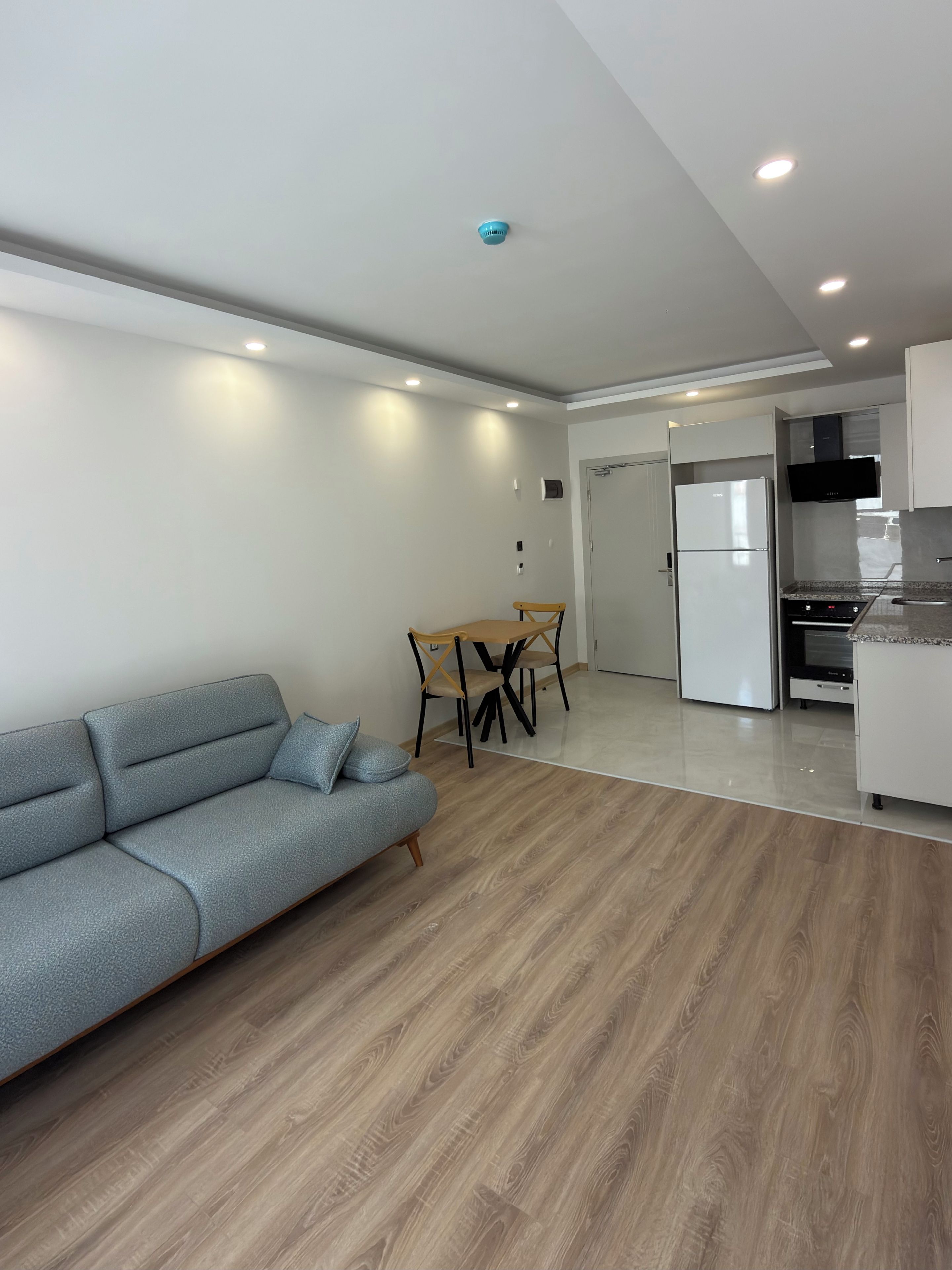 Una sala moderna con un sofá gris, una mesa de comedor de madera y sillas. Se ve una cocina con armarios blancos y un horno empotrado al fondo.