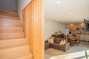 Un interior moderno con una escalera de madera, una sala de estar con sofá y cojines, una mesa de comedor y una cocina con armarios de madera.