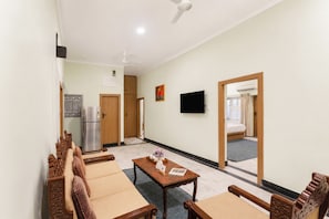 A living room with a sofa set, a coffee table with decorative items, a flat-screen TV mounted on the wall, and a kitchen area with a refrigerator and cabinets. - Alaya Stays Aira Amar Villa 3BHK (Udaipur)