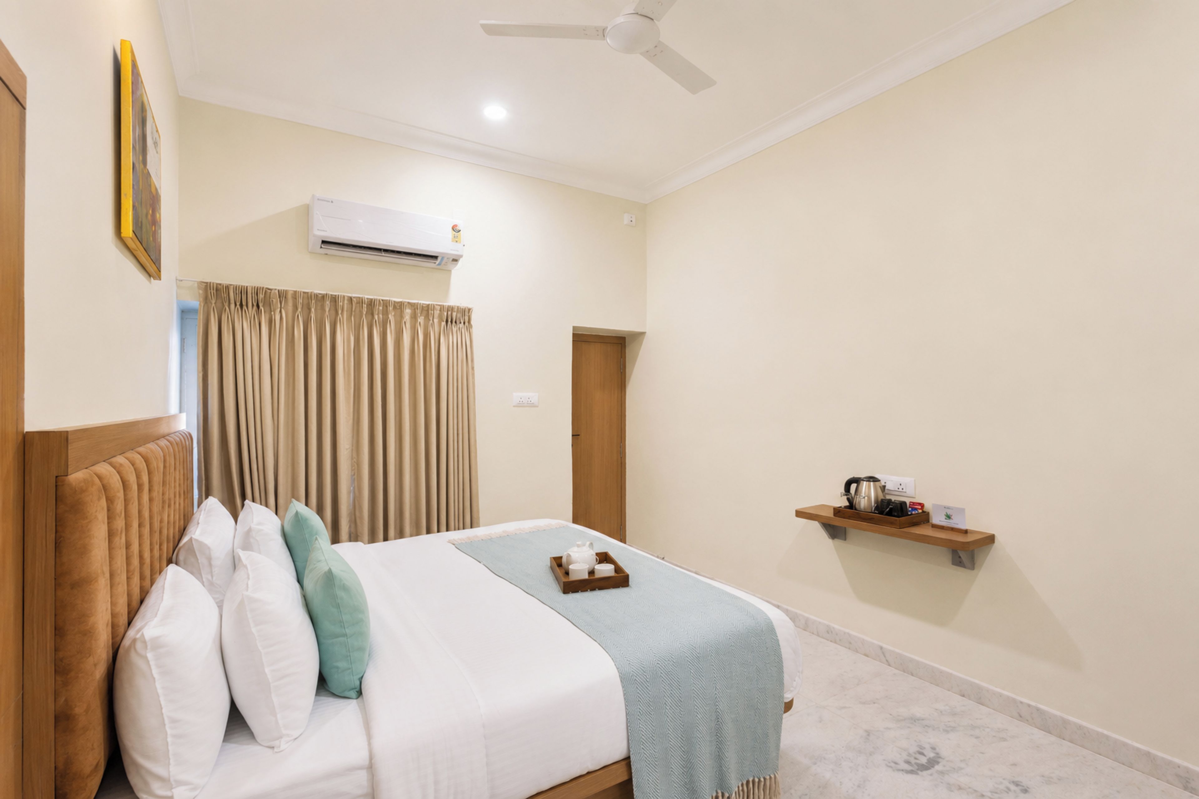 Habitación de hotel con cama, ventilador de techo, aire acondicionado, ventana con cortinas y una pequeña estantería con un hervidor y un cartel.