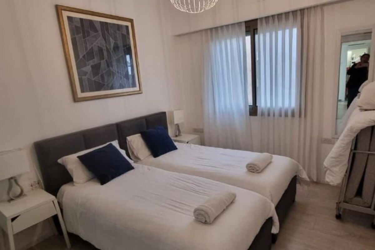 A hotel room with two beds, a nightstand, a lamp, a framed picture on the wall, and a window with curtains.
