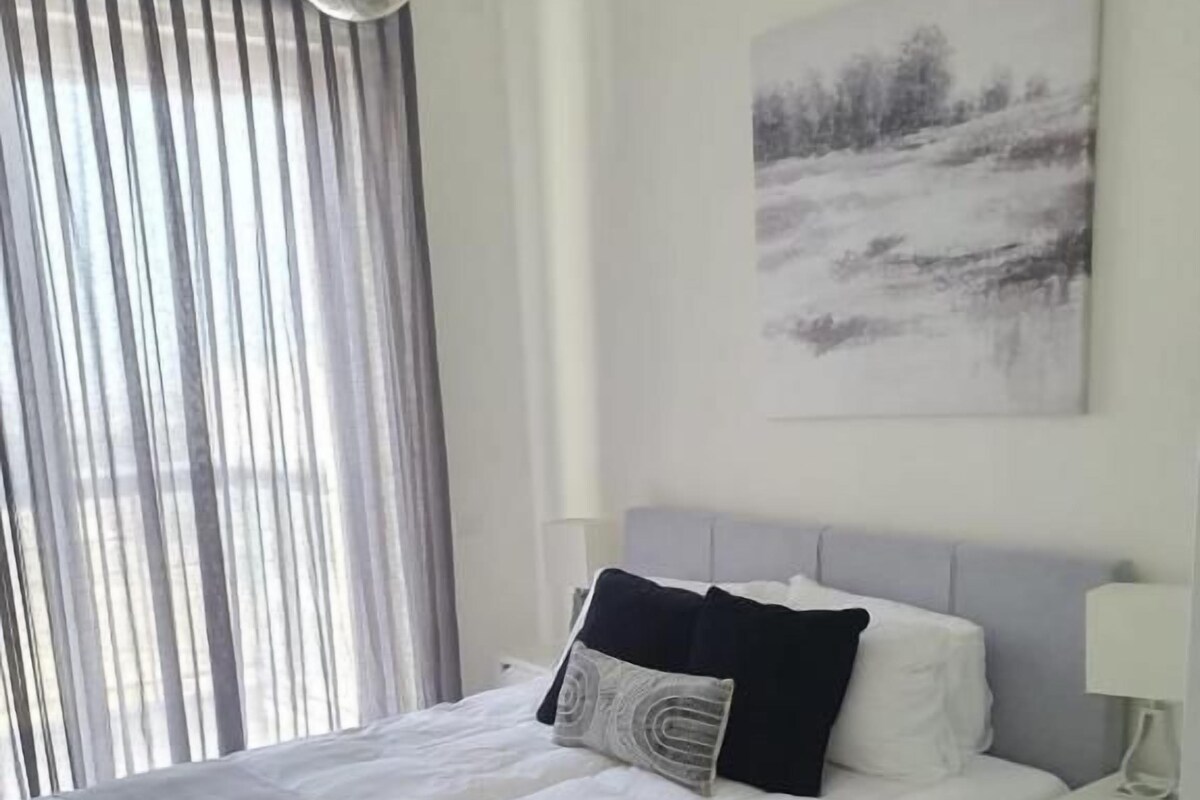A neatly made bed with a grey headboard, a bedside table with a lamp, and a painting on the wall.