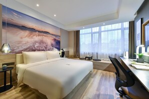 Habitación de hotel con una cama grande, un escritorio con una silla, una lámpara de escritorio, una mesita con una planta y un ventanal con cortinas.