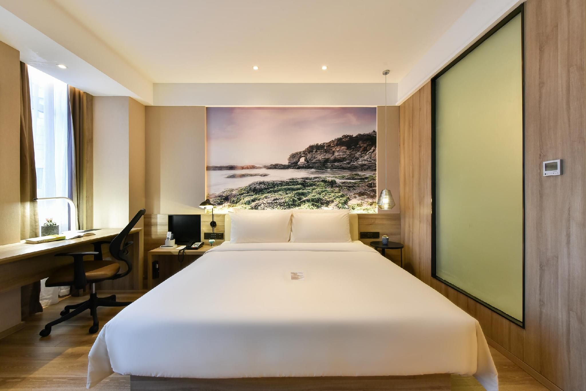 A hotel room with a large bed, a desk, a chair, a nightstand, a lamp, and a view of a coastal landscape.