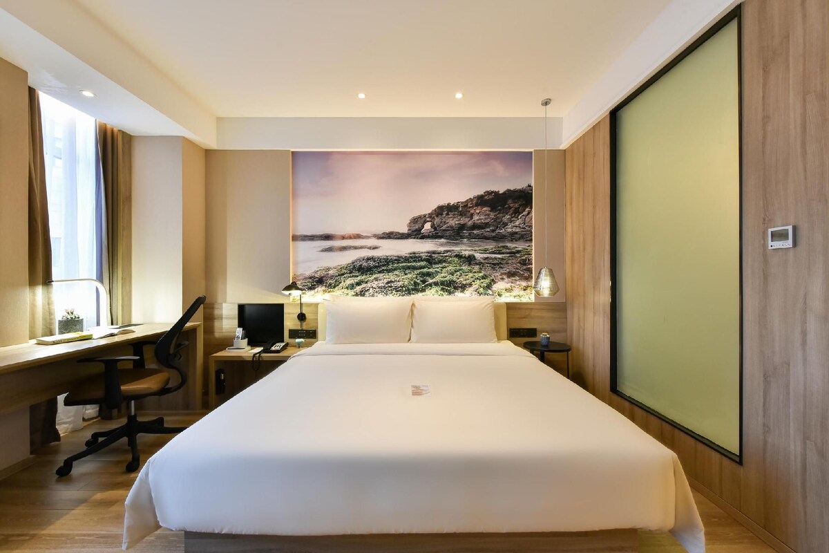 A hotel room with a large bed, a desk, a chair, a nightstand, a lamp, and a view of a coastal landscape.