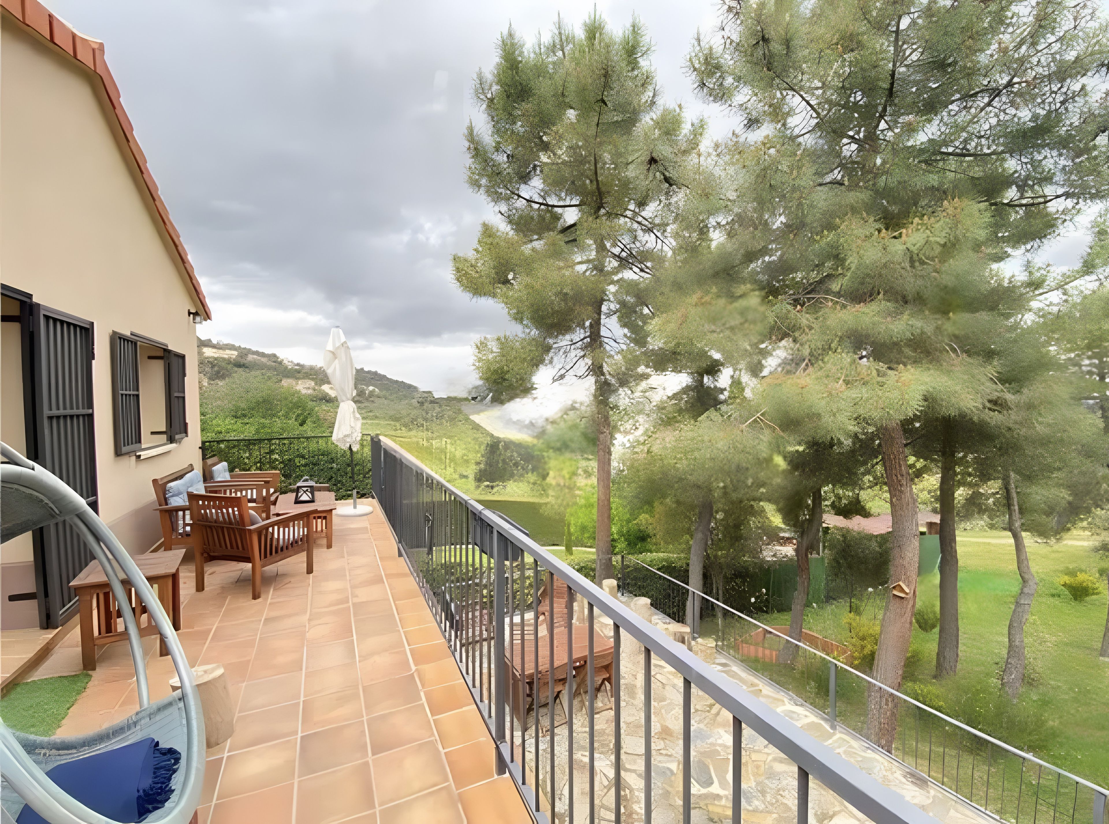 A balcony with a view of a green landscape, a wooden table, and a hanging chair.