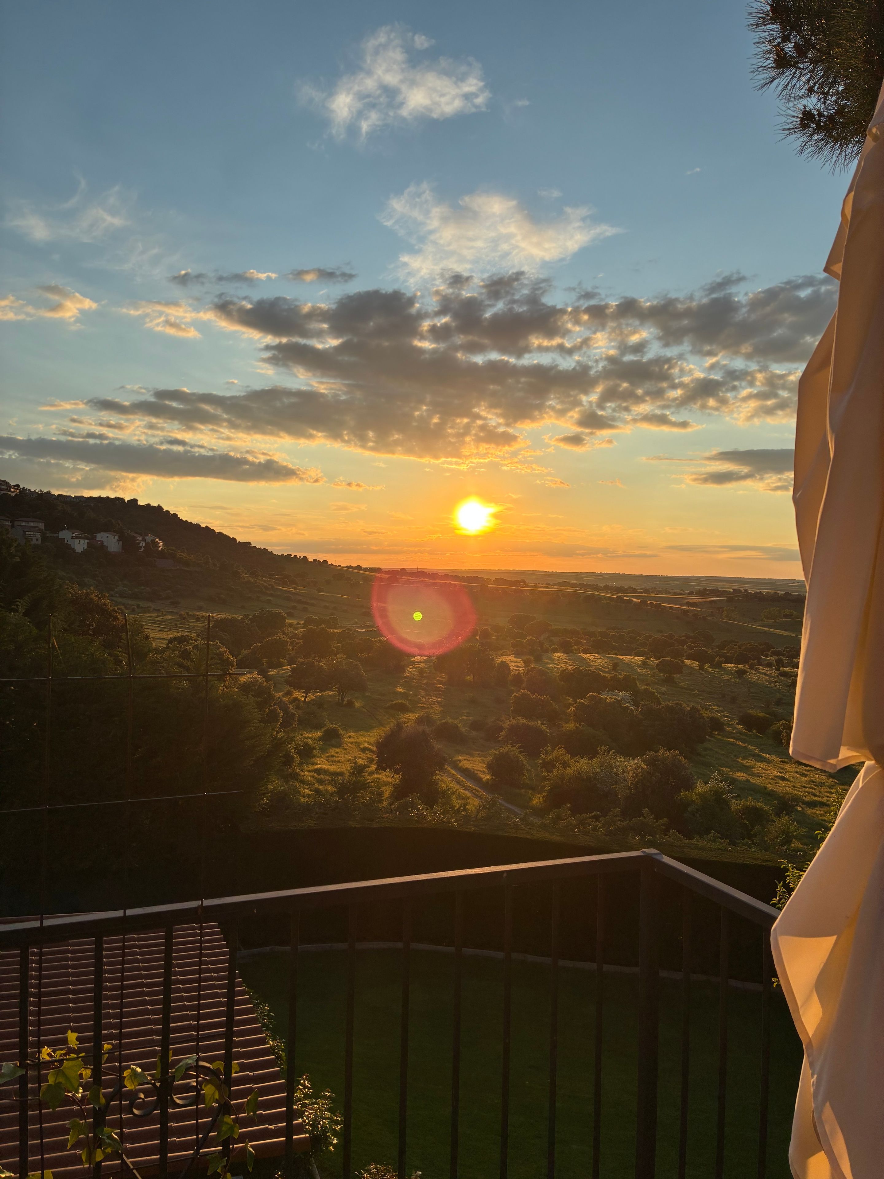Vista del atardecer desde un balcón con barandilla, con vistas a un paisaje montañoso con árboles dispersos y un cielo despejado.