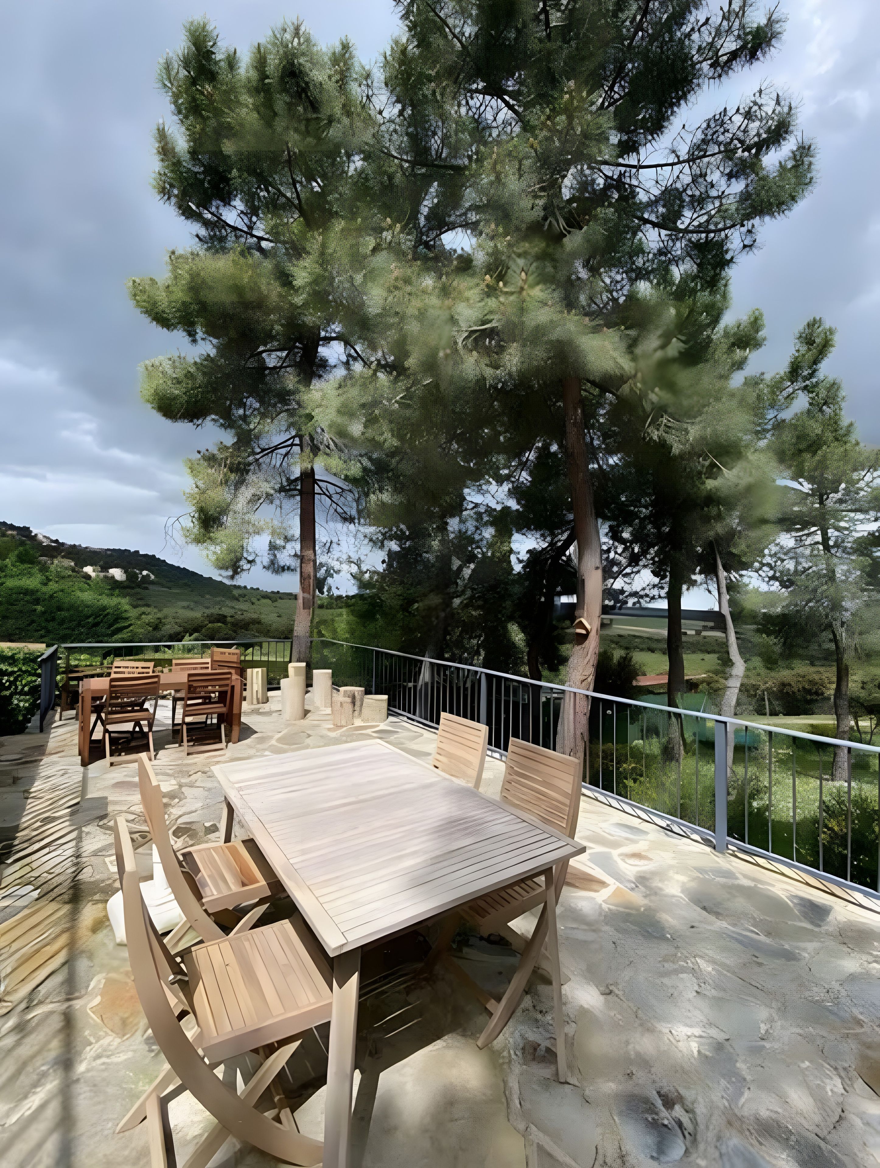 Un patio al aire libre con mesa y sillas de madera, rodeado de piso de piedra y barandilla de metal.