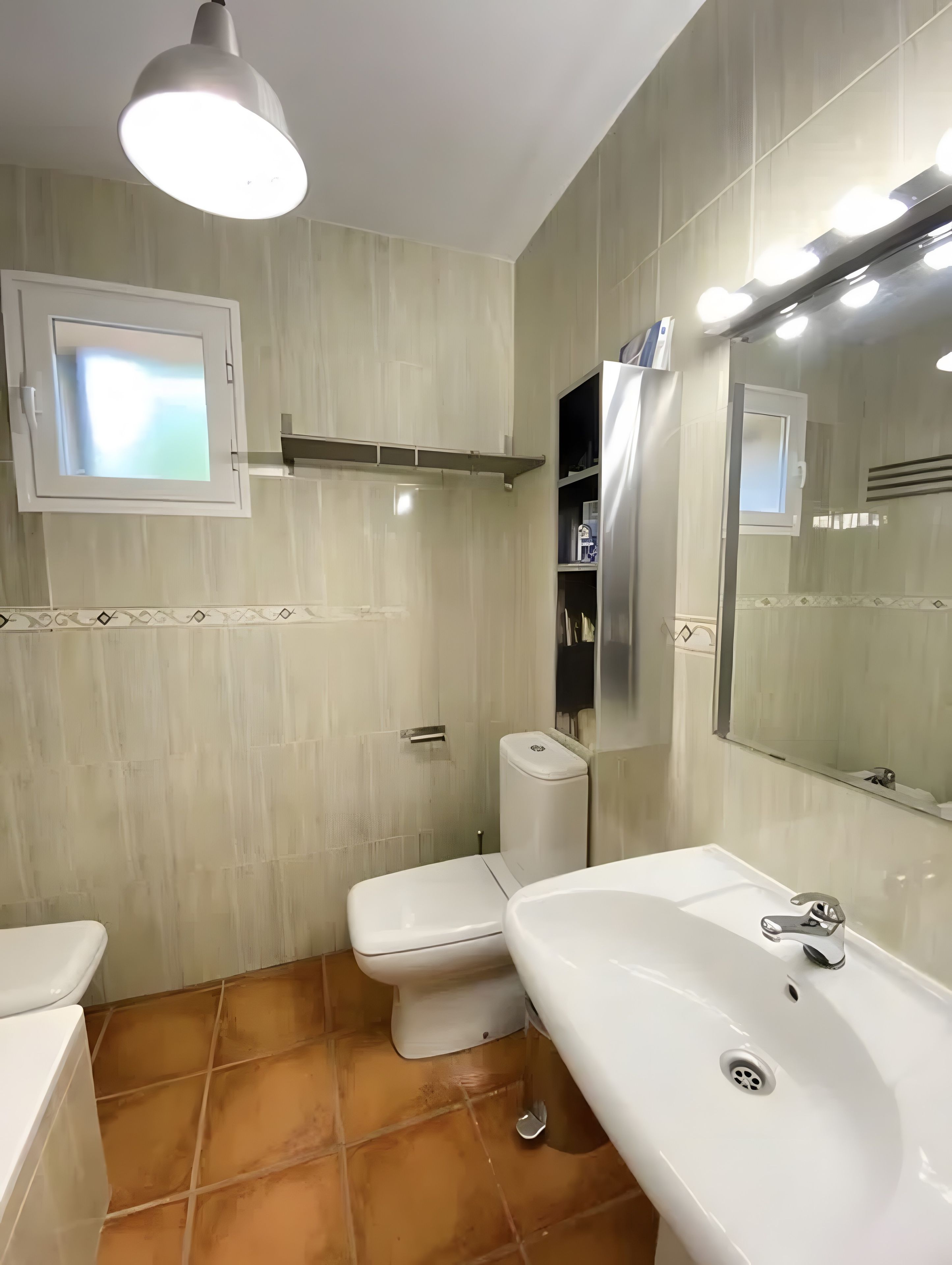 Une salle de bain avec des toilettes, un lavabo et un miroir. Il y a une fenêtre au-dessus des toilettes et une étagère au-dessus du lavabo.
