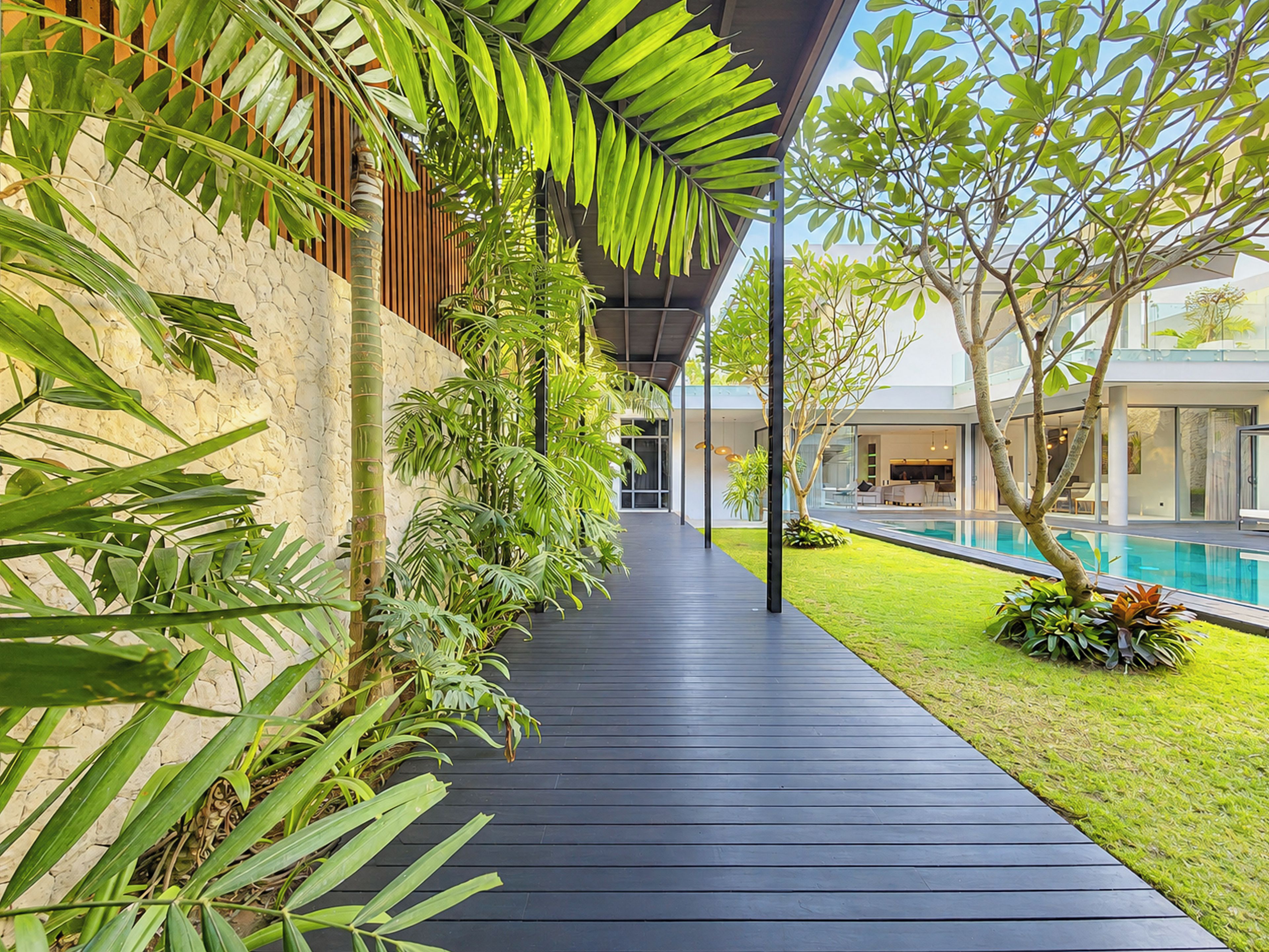 Un paseo techado junto a una piscina, rodeado de exuberante vegetación y un edificio moderno.