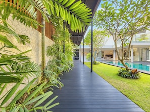 A covered walkway with a poolside area, surrounded by lush greenery and a modern building. - Cyrille Villa Jimbaran by Nagisa Bali (Badung)