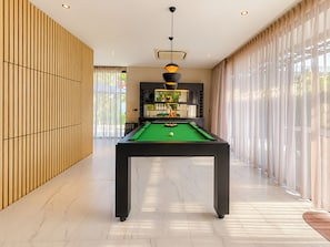 Une table de billard dans une pièce bien éclairée, au design moderne, avec de grandes fenêtres et une vue sur la verdure à l’extérieur.