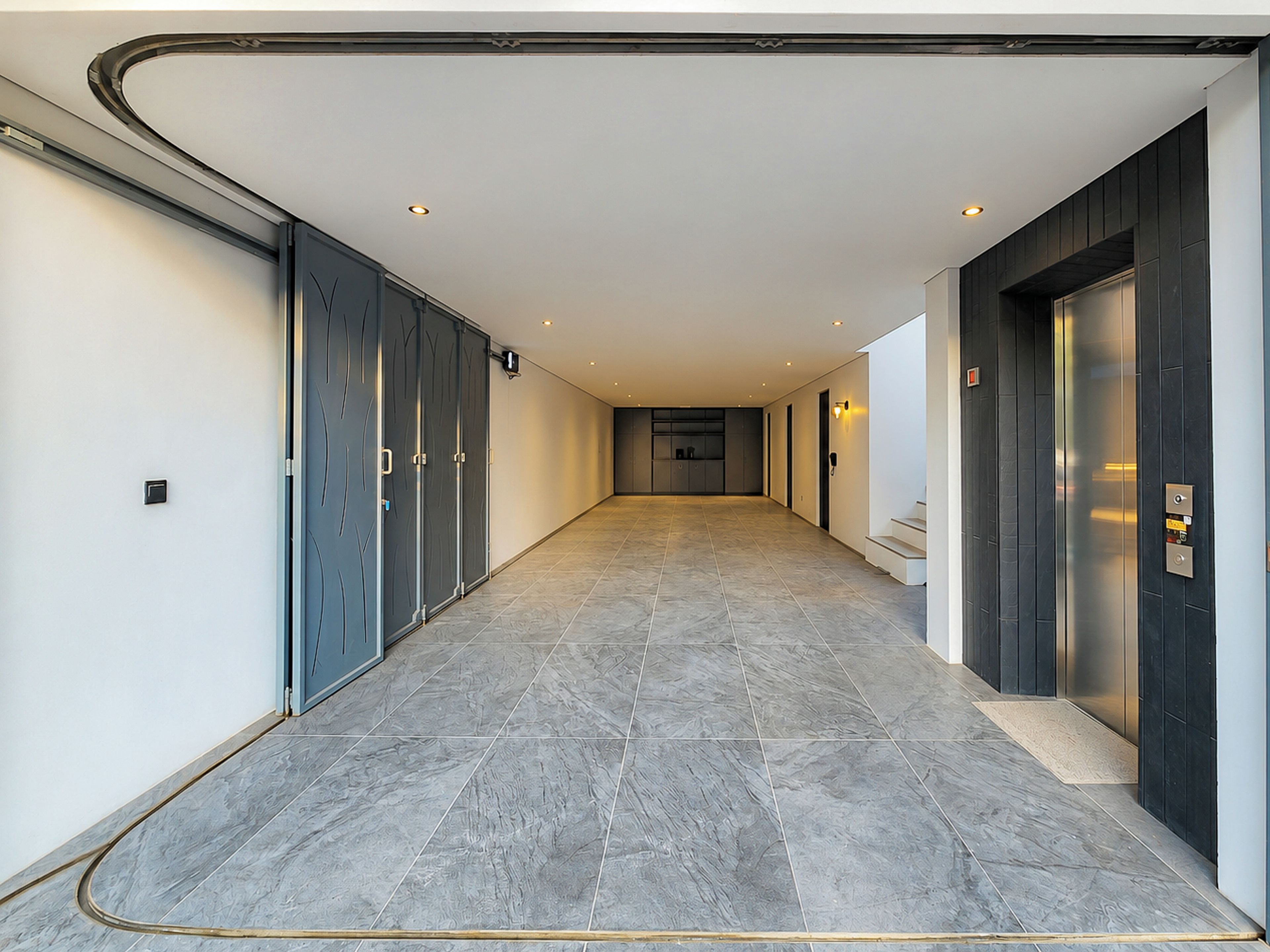 Um corredor com piso de azulejos, uma porta com teclado numérico e um aparelho de luz fixado na parede.