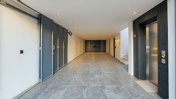 Um corredor com piso de azulejos, uma porta com teclado numérico e um aparelho de luz fixado na parede.