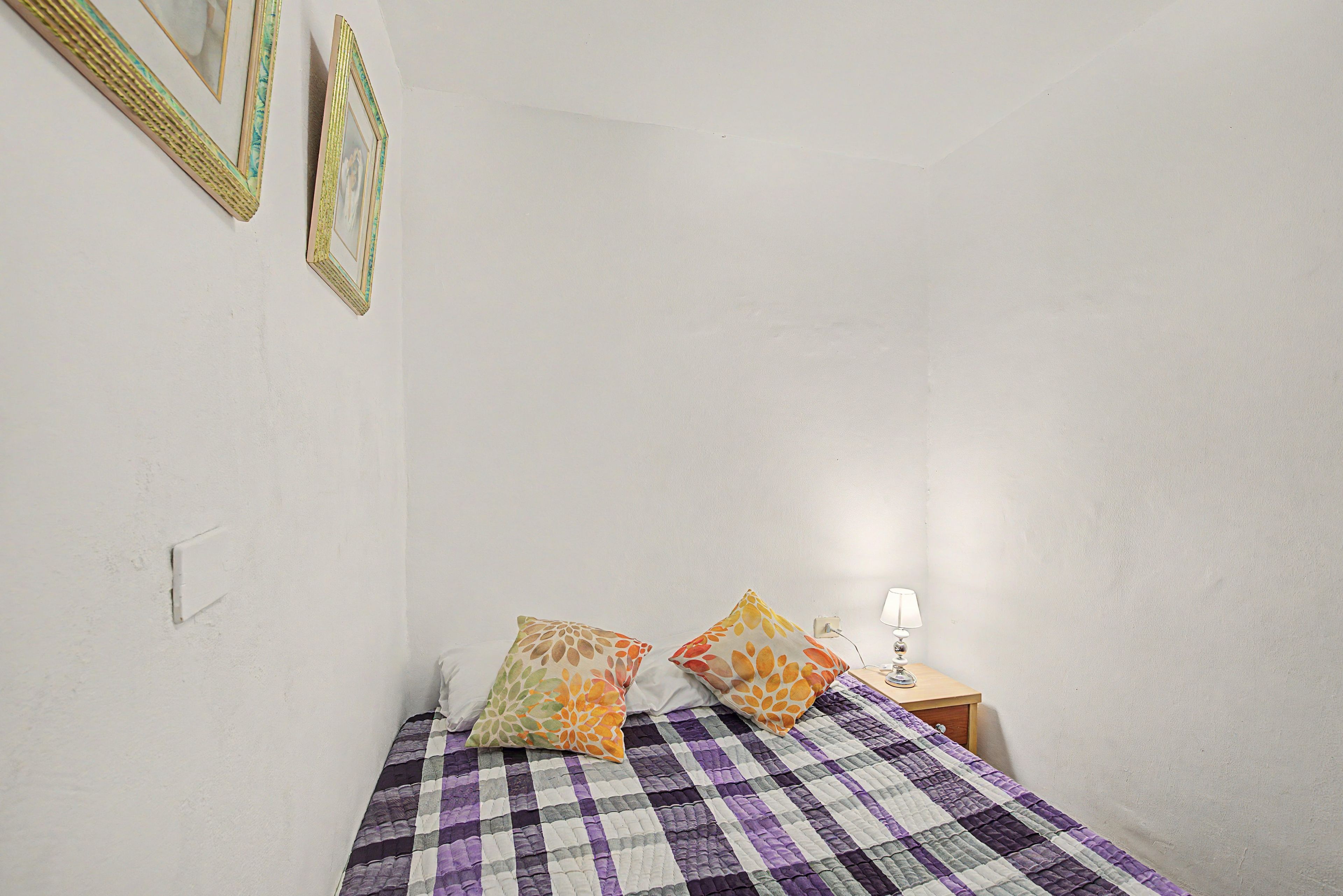 Un dormitorio con una colcha a cuadros, dos almohadas, una mesita de noche con una lámpara y cuadros en la pared.