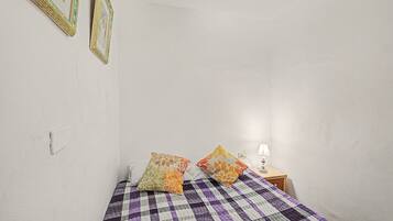 Un dormitorio con una colcha a cuadros, dos almohadas, una mesita de noche con una lámpara y cuadros en la pared.