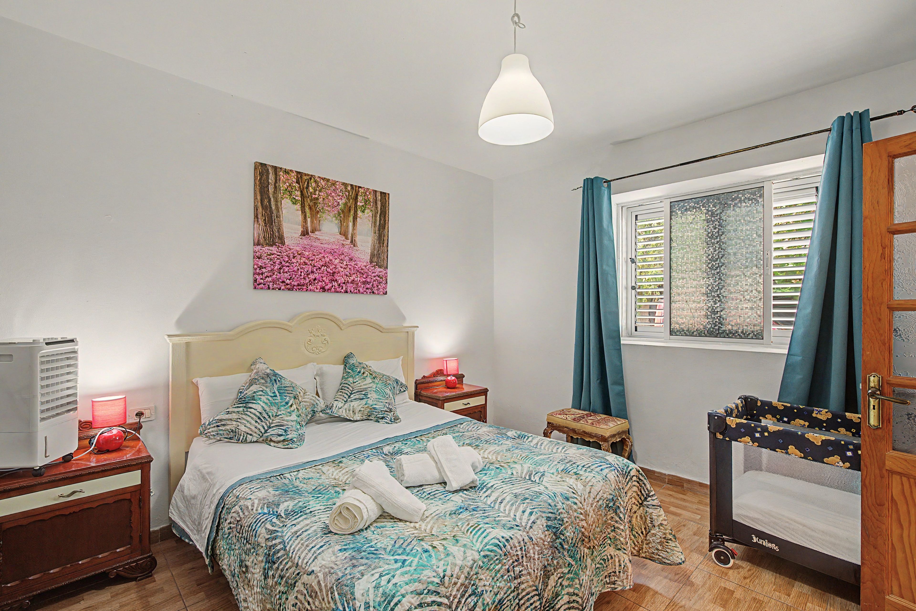 Een slaapkamer met een bed, nachtkastje, raam met gordijnen en een aan de muur bevestigd schilderij.