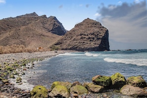 Una playa rocosa con aguas cristalinas, un edificio pequeño y montañas al fondo.