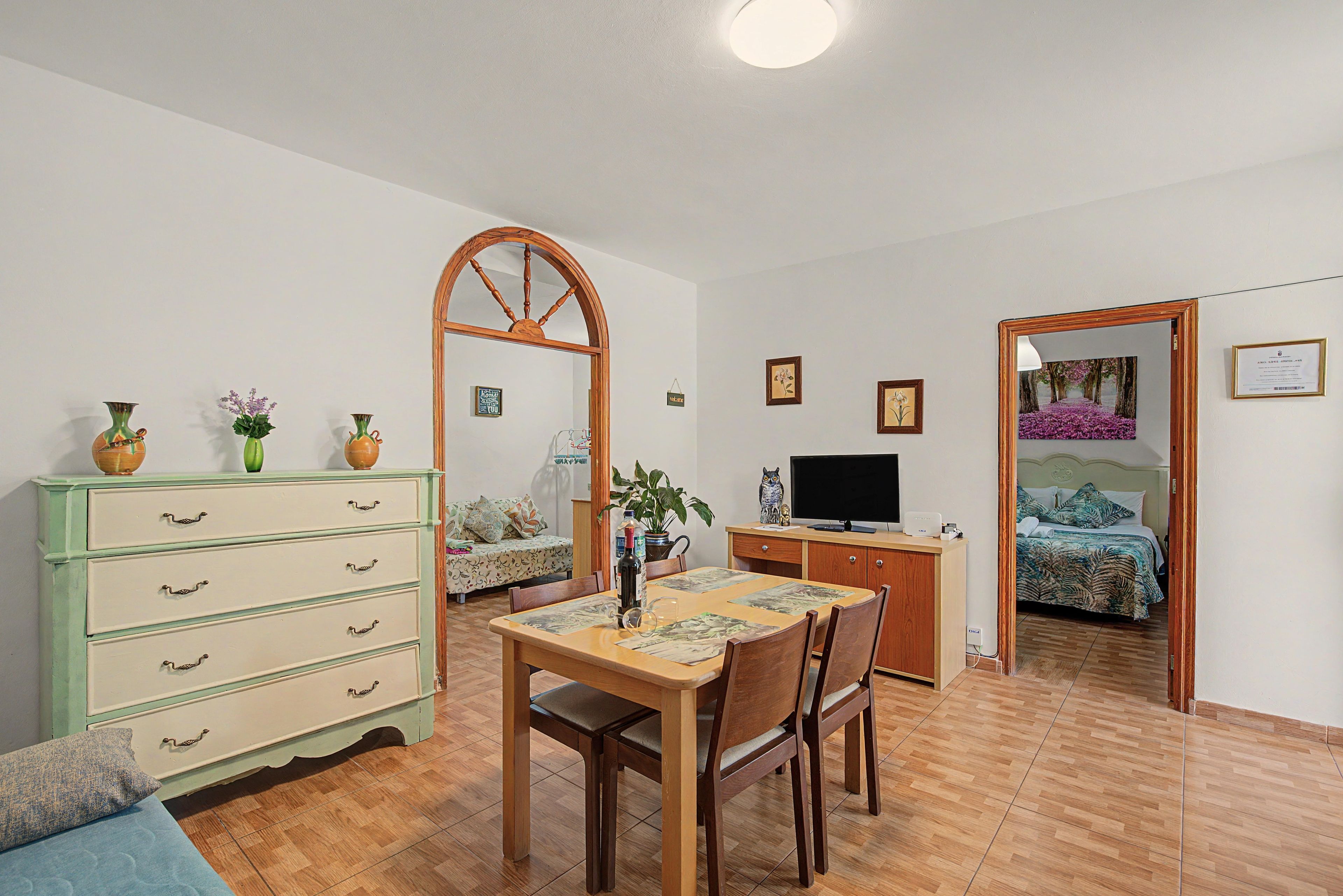 Una habitación con una mesa de comedor, sillas, un aparador de madera, un televisor y una cama con colcha estampada.