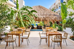 Un espacio de comedor al aire libre con mesas y sillas, una piscina y una estructura con techo de paja.
