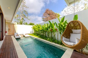 Una piscina con cubierta de madera, una silla colgante y abundante vegetación.