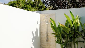 Pared de ducha de piedra con ducha de mano, rodeada de exuberante follaje verde.