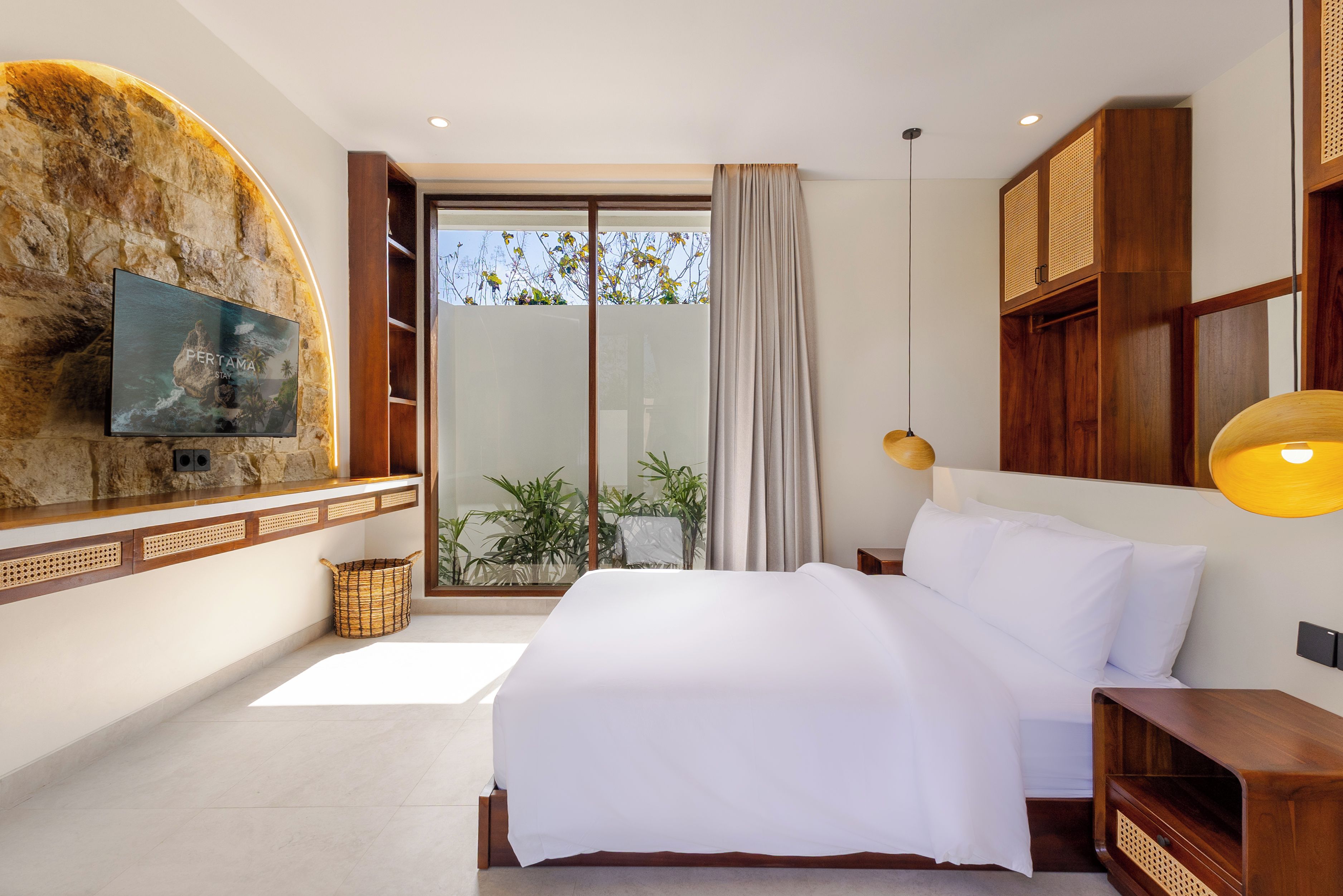 A hotel room with a bed, a TV mounted on a stone wall, a large window with a view of trees, and a wooden wardrobe.