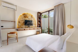 A hotel room with a bed, a desk, a chair, a stone wall with a picture, a bookshelf, and a window with curtains. - Niscaya Villa Uluwatu by Pertama Stay (Badung)