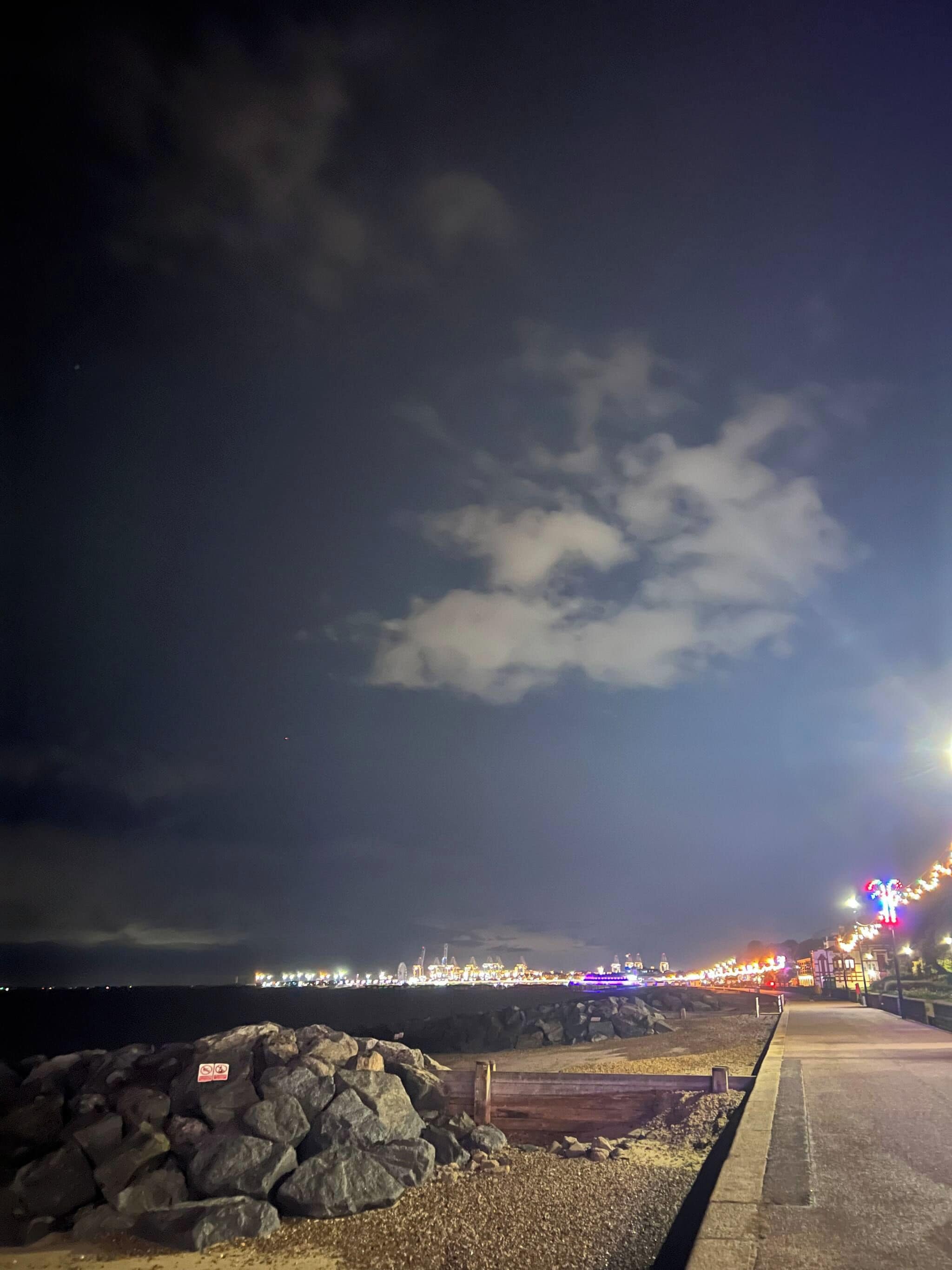 A nighttime coastal scene with a rocky shoreline, a paved walkway, and a distant illuminated pier.