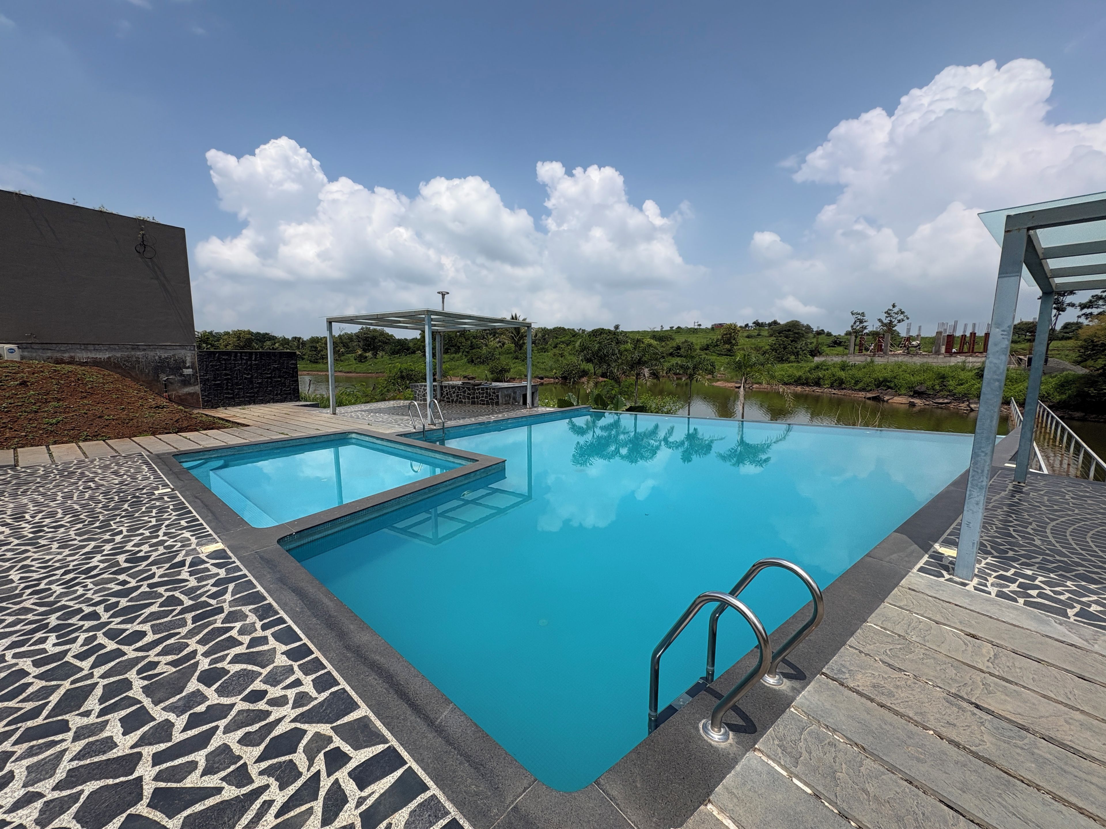 A rectangular swimming pool with a stone-patterned deck, a covered seating area, and a view of a natural landscape.